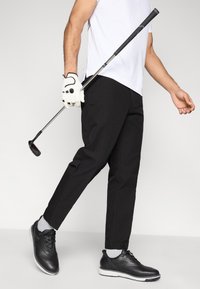 Μαύρο παντελόνι για γκολφ με στενή εφαρμογή, σε συνδυασμό με ασπρόμαυρα παπούτσια. Το άτομο κρατάει ένα γκολφ κλαμπ, φοράει λευκό πουκάμισο γκολφ και γάντι.