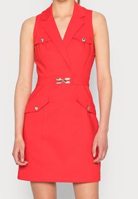 Sleeveless red dress with lapel collar, four flap pockets with gold buttons, and a belt with a central gold buckle.