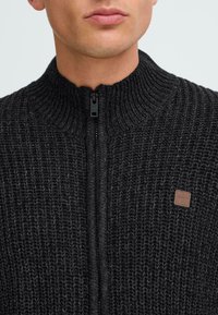 Maglione nero a maglia con colletto alto e cerniera, caratterizzato da un motivo testurizzato e da un piccolo etichetta del marchio marrone sul petto.