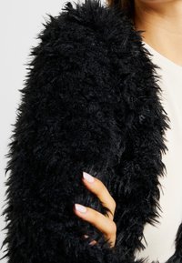Giacca nera fluffy realizzata in un materiale testurizzato simile alla pelliccia; primo piano della manica e della spalla che mette in evidenza la superficie morbida e tattile.