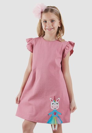 Niña joven con un vestido rosa con mangas con volantes y un parche de unicornio colorido, sonriendo frente a un fondo claro y sencillo.