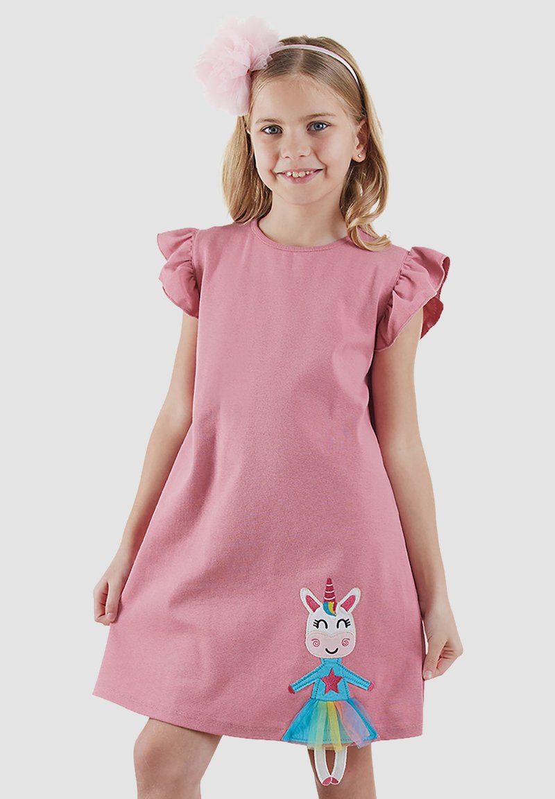 Ung pige iført en pink kjole med flæser på ærmerne og en farverig enhjørningemærke smilende mod en ensfarvet lys baggrund.