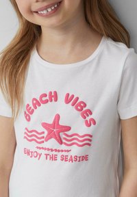 Witte katoenen t-shirt met roze tekst "BEACH VIBES" en "ENJOY THE SEASIDE", met een roze zeester grafiek en golvende lijnen als accenten.