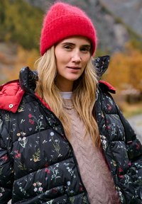 Čierna nadýchaná bunda s kvetinovým vzorom, červenými akcentmi a kontrastnou červenou pletenou čiapkou. Pod ňou texturovaný béžový sveter.