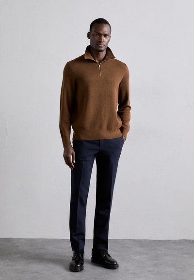 Pull en tricot marron avec un col montant et une fermeture éclair argentée, associé à un pantalon tailleur bleu marine foncé et des chaussures à lacets noires.