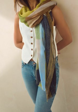 Femme portant un gilet sans manches blanc boutonné, un jean bleu et une écharpe multicolore drapée autour du cou et des épaules.