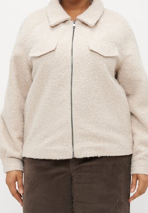 Persona con chaqueta beige claro texturizada con cremallera y bolsillos de solapa en el pecho, y pantalones de pana marrón oscuro.