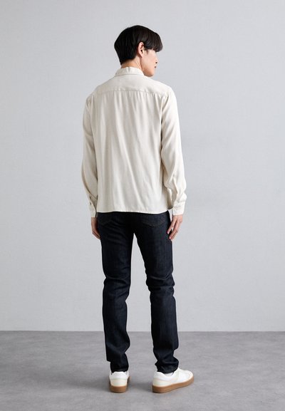 Lichtbeige langemouwenshirt met knoopsluiting, gecombineerd met donkerblauwe jeans en witte sneakers, gezien vanuit de achterkant.