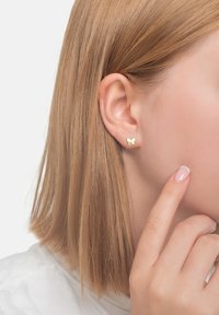 Goldene, schmetterlingsförmige Ohrstecker an dem Ohr eines Modells; glatte, metallische Oberfläche; kleine Größe; nah am Ohrläppchen platziert.