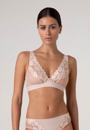 Donna con capelli scuri che indossa un reggiseno in pizzo rosa pallido e mutandine abbinate su sfondo chiaro uniforme.