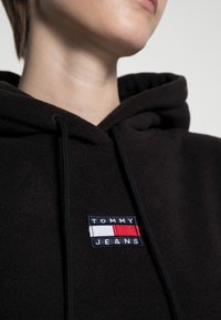 Felpa nera con cappuccio regolabile. Presenta un patch ricamato blu, bianco e rosso con la scritta "TOMMY JEANS." Texture liscia.