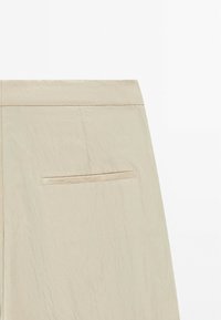Section d'une jupe en cuir beige montrant une poche passepoilée arrière avec une couture propre sur fond blanc uni.