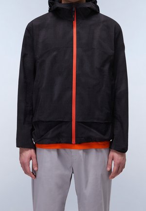 Homme portant une veste noire à capuche avec une fermeture éclair rouge sur une chemise orange et un pantalon gris clair sur fond uni.