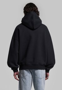 Felpa nera oversize con un grande cappuccio, spalle scese e polsini a coste. Tessuto morbido, indossata con jeans denim azzurri chiari.