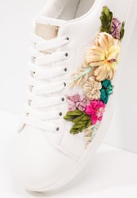 Bílé kožené tenisky s květinovou výšivkou v růžové, zelené, zlaté a modré barvě, s dekorativními prvky a bílými tkaničkami.