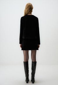 Top lungo in velluto nero abbinato a una mini gonna coordinata, completato da stivali neri in pelle al ginocchio e collant trasparenti.