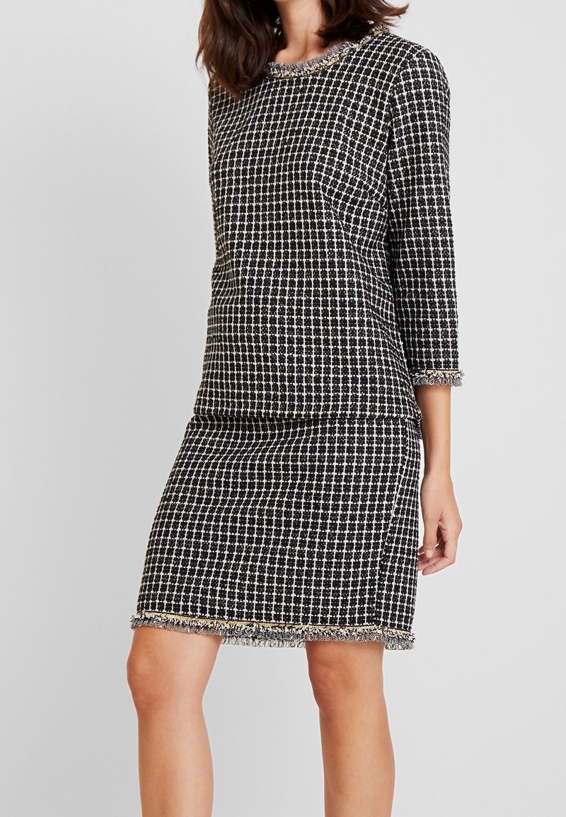 Femme portant une robe à carreaux noirs et blancs avec des manches trois-quarts et un bord à franges aux manches et à l'ourlet, sur un fond uni.