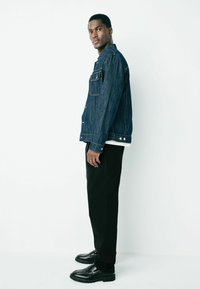 Denim jacket in dark blue with pockets and silver buttons, paired with black trousers and shiny black loafers. Model stands sideways.