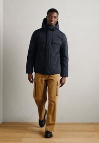 Casaco puffer preto com capuz, duas bolsos no peito, usado com calças de carga bege. O modelo está de pé sobre um piso de madeira contra uma parede neutra.