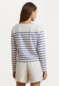 Haut à manches longues blanc avec des rayures horizontales bleues, présentant un décolleté décontracté et une coupe légèrement raccourcie. Porté avec un short de couleur claire.