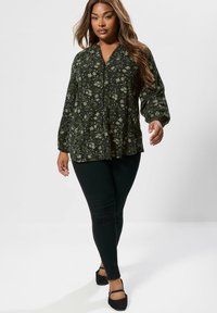 Dunkelgrüne Bluse mit floralem Muster, lockerer Passform, V-Ausschnitt und Puffärmeln. Kombiniert mit schwarzen, figurbetonten Hosen und stylischen schwarzen Schuhen.