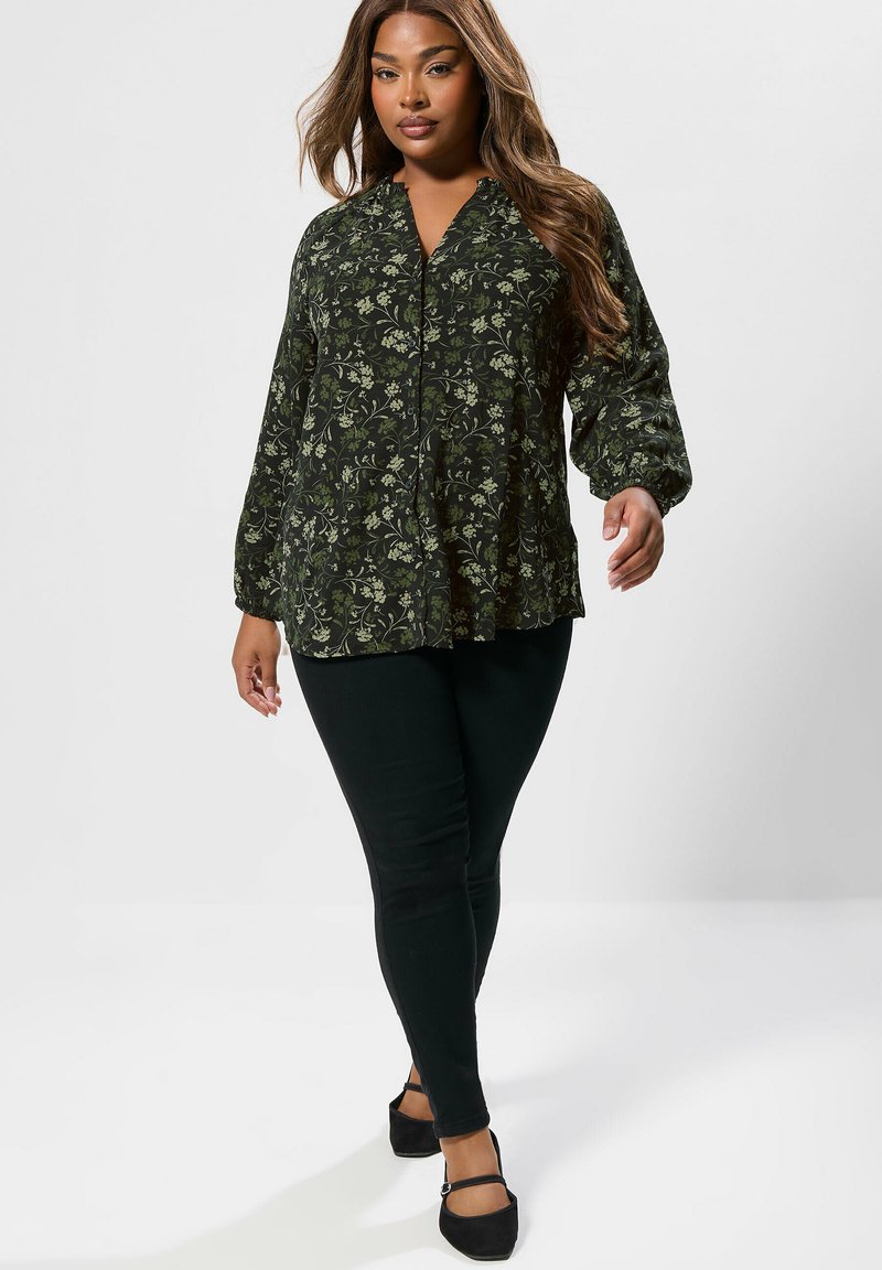 Dunkelgrüne Bluse mit floralem Muster, lockerer Passform, V-Ausschnitt und Puffärmeln. Kombiniert mit schwarzen, figurbetonten Hosen und stylischen schwarzen Schuhen.