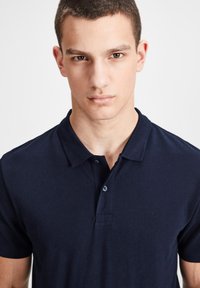 Polo shirt bleu marine en coton texturé avec col, patte de boutonnage à trois boutons et un logo subtil sur la poitrine. Manches courtes.