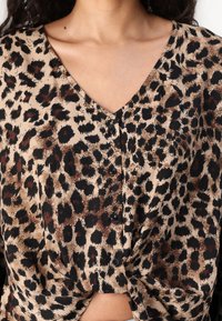 Bluse med leopardmønster og V-hals, med sorte knapper og knude-design i taljen. Lavet af letvægtsstof med en glat tekstur.