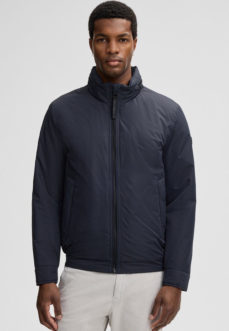 Donkerblauwe gewatteerde jas met een hoge kraag, ritssluiting aan de voorkant, zijzakken en een gladde, matte textuur. Met elastische manchetten.