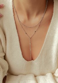 Gelaagde zilveren kettingen met fijne schakels, waarvan er één kleine kralen en een hangende medaille heeft, gedragen over een crèmekleurige gebreide trui.