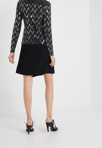 Svart paljett långärmad topp med zigzag mönster, tillsammans med en svart A-linjeformad kjol. Modellen bär svarta klacksandaler med bakremmar.