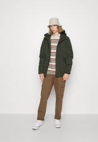Olivengrønn polstret jakke med hette, båret over en beige og stripete skjorte, kombinert med brune cargo-bukser og hvite sneakers.
