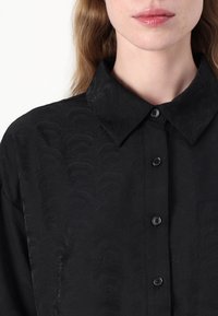 Camisa preta de botões com um padrão de ondas subtil, com colarinho, seis botões e um bolso de chapa no lado esquerdo.