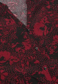 Zwarte stoffen top met een rode bloemen- en stippenpatroon, met een v-halsontwerp en zwarte knopen aan de voorkant. Geweven gelaagd materiaal zichtbaar.