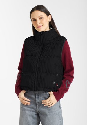 Schwarze gesteppte Weste mit hohem Kragen, gerippter Textur, Frontreißverschluss und seitlichen Knöpfen, kombiniert mit einem roten Pullover mit Herzdetails.
