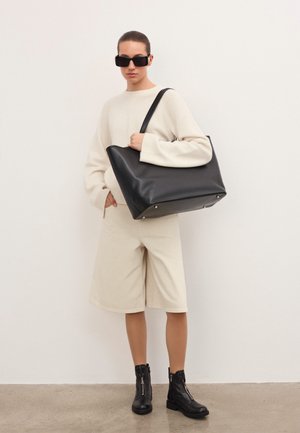 Frau in einem übergroßen cremefarbenen Pullover und Shorts, trägt schwarze Knöchelstiefel und Sonnenbrille, hält eine große schwarze Leder-Tote-Bag auf der Schulter.