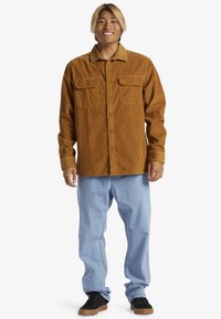 Brun kordfløjlsskjorte med to brystlommer, knaplukning, lange ærmer og en afslappet pasform, parret med lyseblå jeans og sorte sneakers.