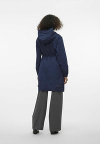 Marineblauwe hoodie met elastische taille, lange mouwen en drukknoopdetails. Draagt over grijze wijde broekspijpen, weergegeven vanuit de achterkant.