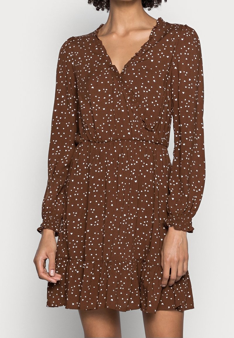 Robe marron à manches longues avec motifs de cœurs blancs, encolure en V, taille élastique, poignets et ourlet à volants, portée par une personne debout.