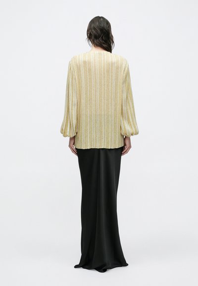 Kvinde med bølget mørkt hår iført en gul- og hvidstribet langærmet top og et langt sort skørt, stående foran en ensfarvet hvid baggrund.