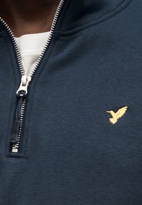 Μπλε ναυτικό φούτερ με φερμουάρ, με υφή, που διαθέτει ασημένιο φερμουάρ και μικρό κίτρινο κεντημένο λογότυπο πουλιού στο στήθος.