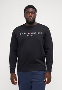 Black sweatshirt made of cotton with a round neck, featuring a white logo text “TOMMY HILFIGER EST. 1985” across the chest.