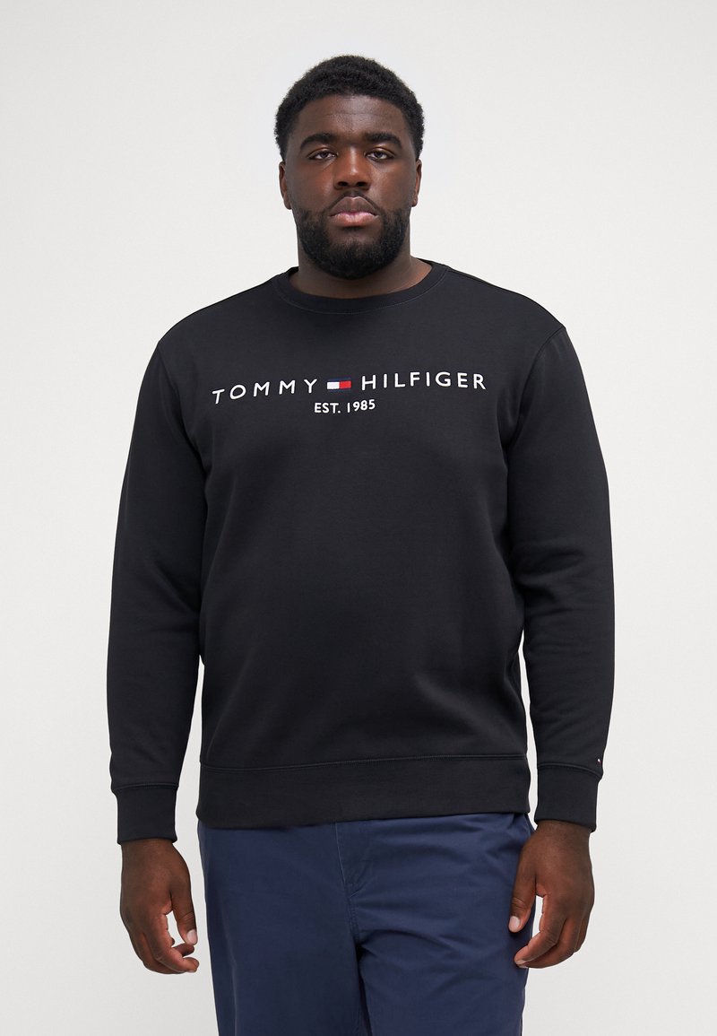 Black sweatshirt made of cotton with a round neck, featuring a white logo text “TOMMY HILFIGER EST. 1985” across the chest.