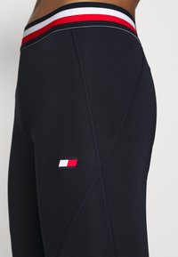 Granatowe legginsy mają wielokolorowy pas w czerwonym, białym i czarnym kolorze, z małą naszywką z logo w kolorze czerwonym, białym i niebieskim.