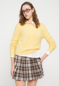 Pull en tricot jaune associé à une chemise blanche à col et une jupe plissée à carreaux marron. La tenue présente une texture douce et un design superposé.