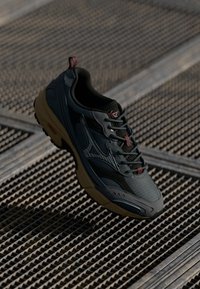 Pantof cu un design sportiv, de culoare neagră și gri, cu talpa bej, așezat pe o suprafață din grilă metalică, evidențiind detaliile de design și materialele texturate.