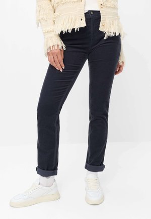 Persoon die donkere opgerolde broek, witte sokken, witte sneakers en een crèmekleurige cardigan met franjes draagt, staand tegen een effen witte achtergrond.