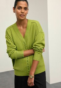 Grüne Strickjacke mit V-Ausschnitt, durchgehender Knopfleiste und gerippten Bündchen. Weiche Textur, lockere Passform und eine aufgesetzte Tasche auf der Vorderseite.