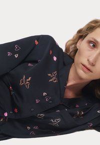 Marineblauw overhemd met kleurrijke patronen van hartjes en lippen, knoopsluiting, kraag en detail van een voorzak. Glad stof met een zachte textuur.