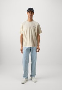 Tricou din bumbac bej cu un mic logo triunghiular pe piept, asortat cu blugi din denim decolorat, de un albastru deschis, și adidași albi, purtat cu o șapcă neagră.
